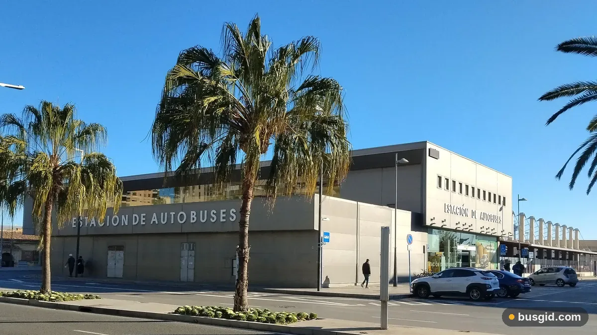 Современное серое здание автовокзала Аликанте с надписью Estación de Autobuses, перед входом пальмы и широкий тротуар